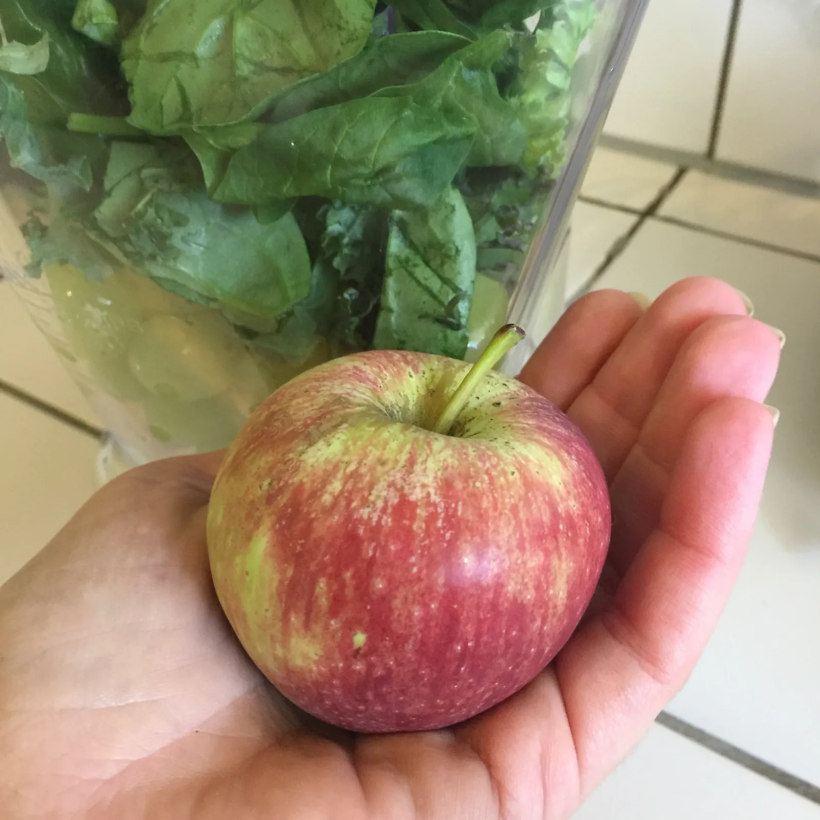 Red and yellow apple held in a hand in front of a blender filled with greens.