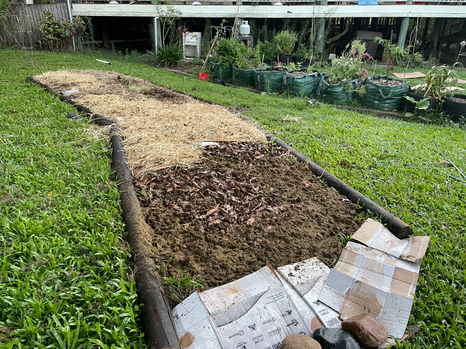 A garden bed framed with logs, featuring sections of straw mulch, soil, and cardboard layers.
